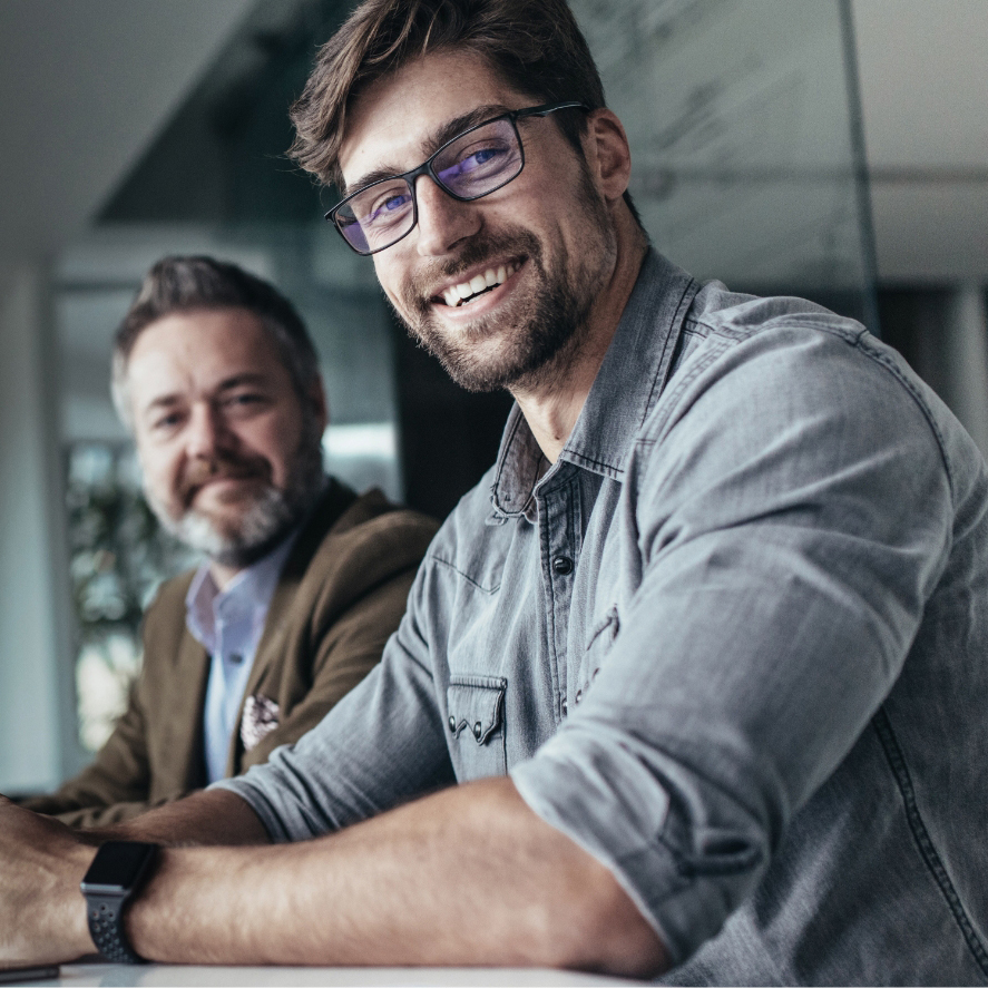Ein lächelnder Mann mit Brille sitzt im Vordergrund eines modernen Büros, während ein Kollege im Hintergrund arbeitet.